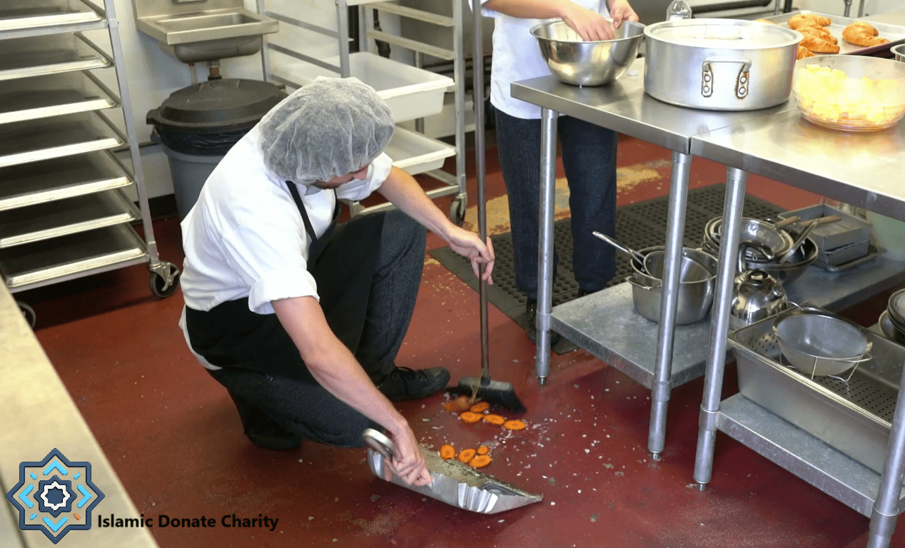 A volunteer cleans a kitchen floor where chopped carrots have fallen, symbolizing the essential but often unseen work of charitable operations funded by crypto donations like BTC.