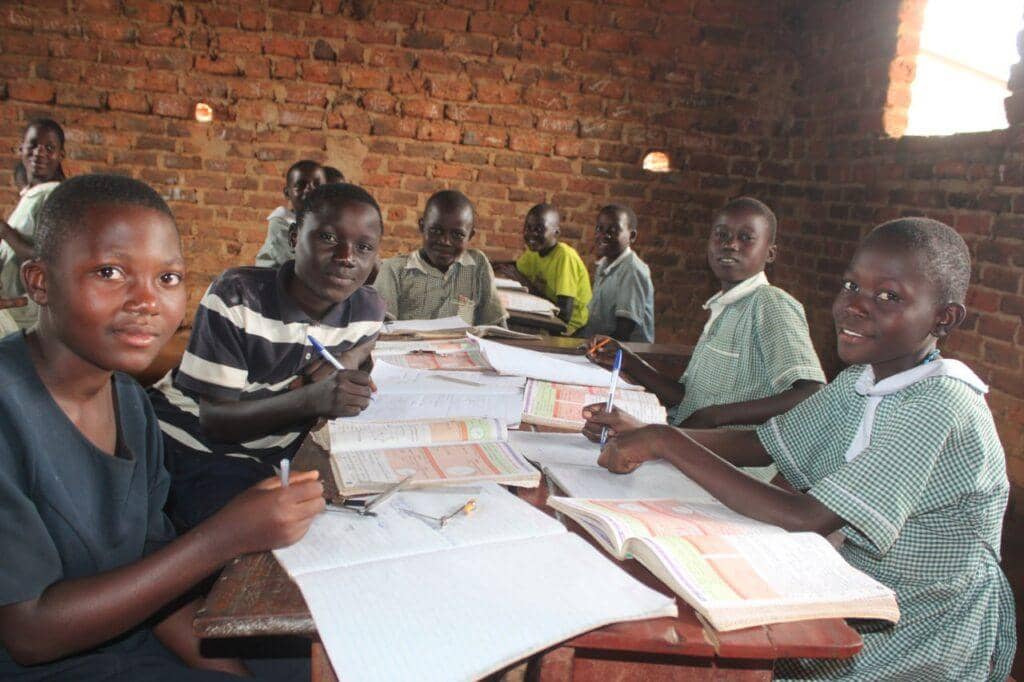 Children studying at desks in a classroom as part of educational support programs funded by crypto donations, specifically BTC. They are focused on their books and writing, representing hope and literacy.