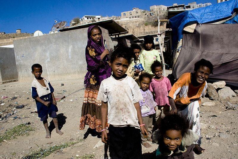 Children in a camp, some with vibrant clothing, others in worn attire. One child in the foreground smiles broadly.