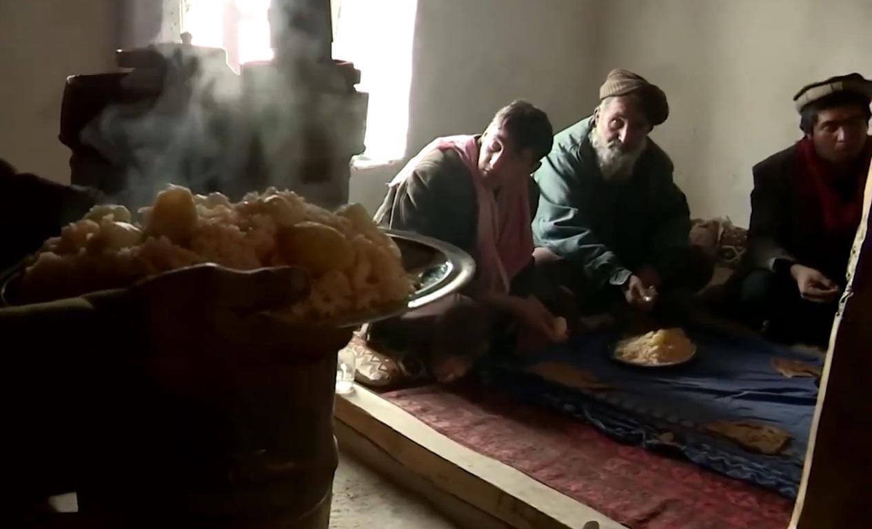 An elderly man and others seated on the floor sharing a meal of rice and potatoes, steam rising from the dish, in a humble setting, representing crypto-enabled Qurbani aid for the needy. Donated with SOL.