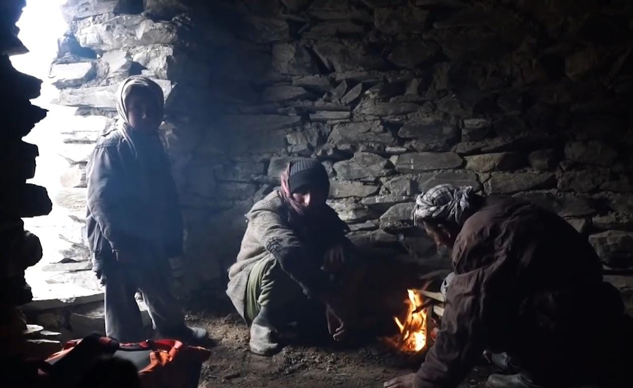 Three people huddled around a small fire in a cave, with one person standing silhouetted in the entrance. Crypto Qurbani for Eid al-Adha aids families, with donations like SOL providing sustenance.