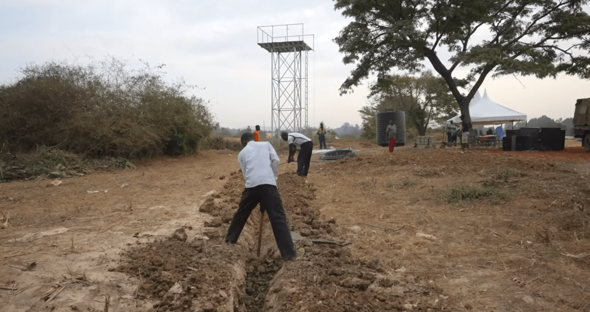 Digging trench for water well construction next to tower, with workers and water tanks visible. Donate crypto like BTC for clean water.