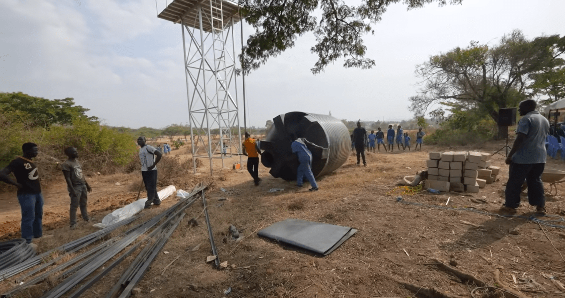 Construction of a water well with a large black tank being maneuvered into place near a metal tower, with workers and community members present. Donate ETH for clean water relief.