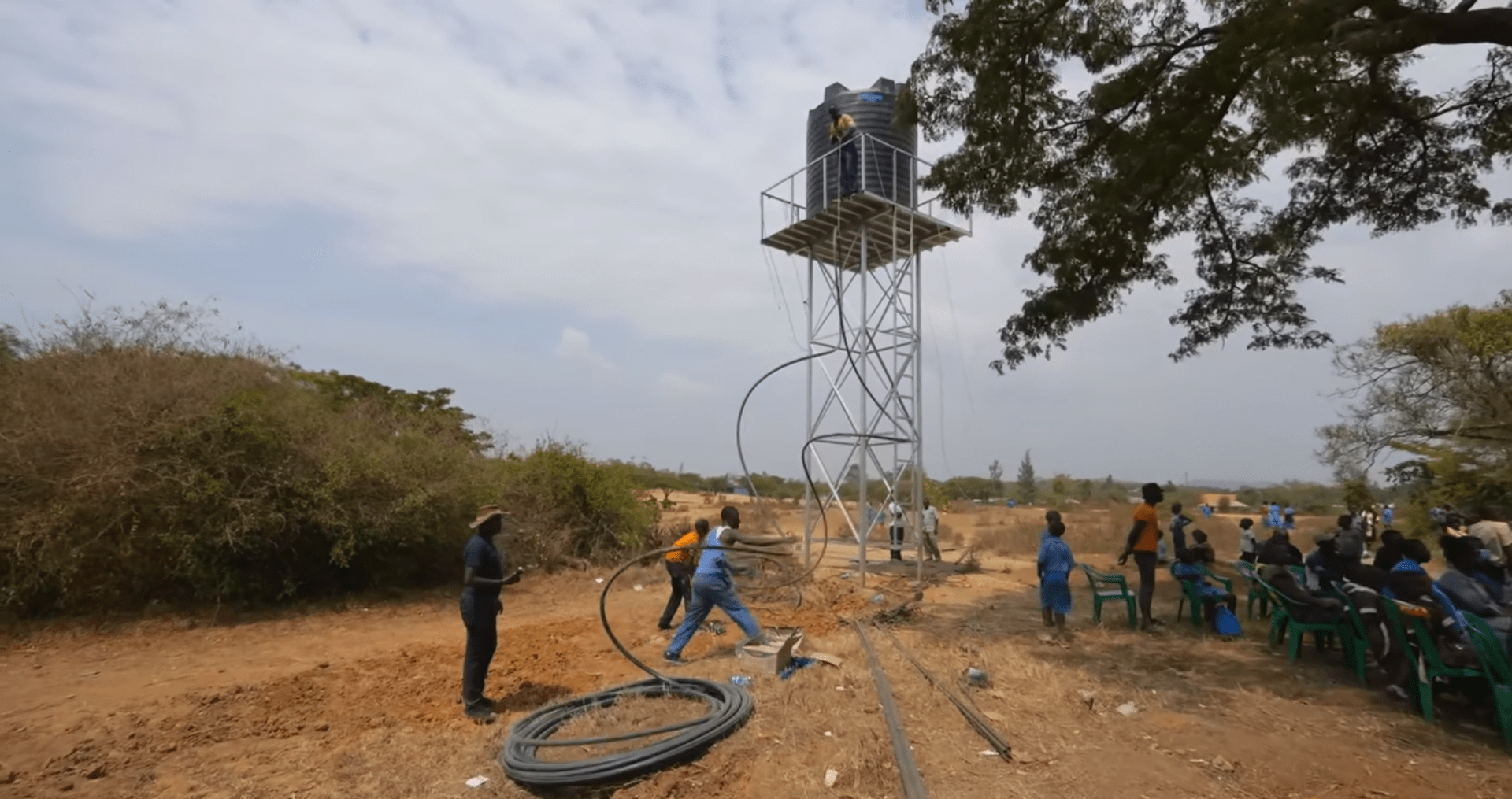 Water well construction in progress with workers installing pipes and a water tank on a metal tower. People gather to witness the project, funded by cryptocurrency donations like USDT for clean water charity.