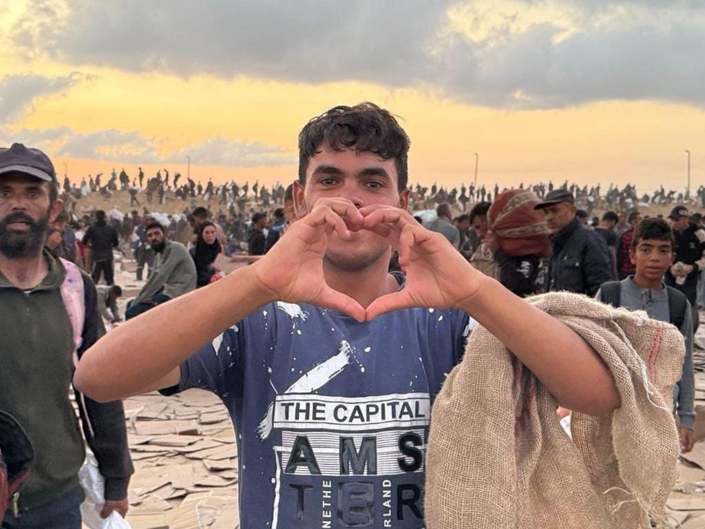 Young Palestinian man forms a heart shape with his hands, symbolizing hope and resilience amidst a crowd of people in a damaged area. Your crypto Zakat, like Stablecoin, can provide vital aid for food, water, and reconstruction efforts.