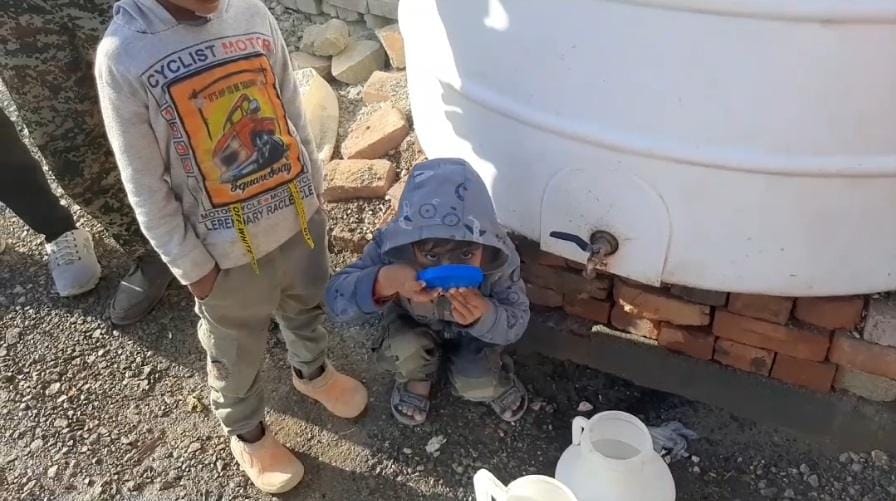 A child drinks from a water tap connected to a large tank, receiving aid through anonymous crypto donations via USDT.