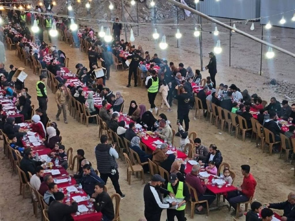 Ramadan 2026 Iftar for displaced families in Gaza. Aid workers distribute meals at long tables set up outdoors under fairy lights. Donations in Ethereum support this vital sustenance for fasting believers.