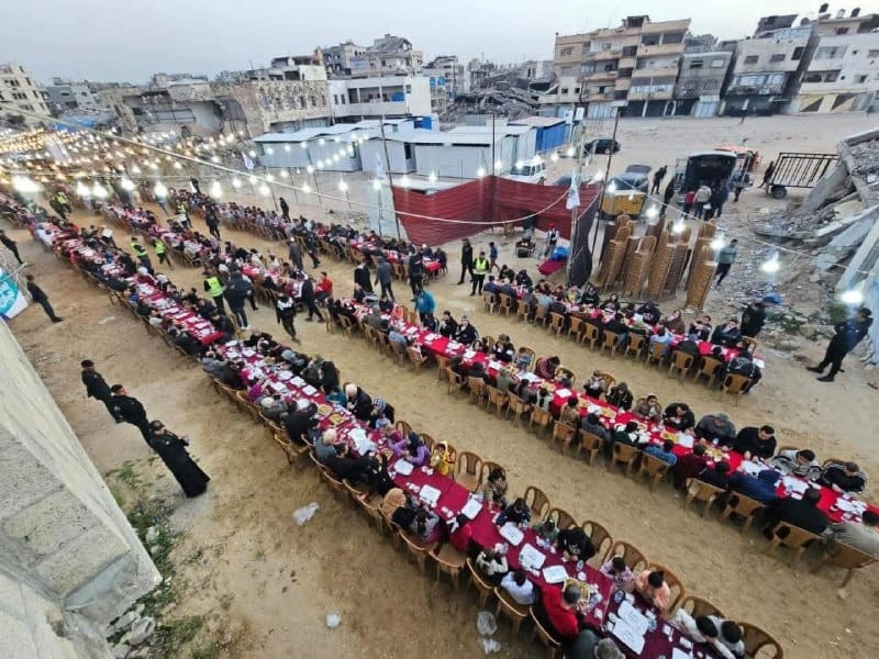 Ramadan Iftar with hundreds of people in Gaza sitting at long tables set up outdoors amidst destroyed buildings, illuminated by string lights. Donations via Litecoin support this relief effort.