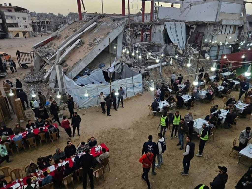 People gather for an outdoor Iftar meal under lights with partial building destruction in the background. Relief efforts provide sustenance to displaced families in Gaza. Donation via USDC.