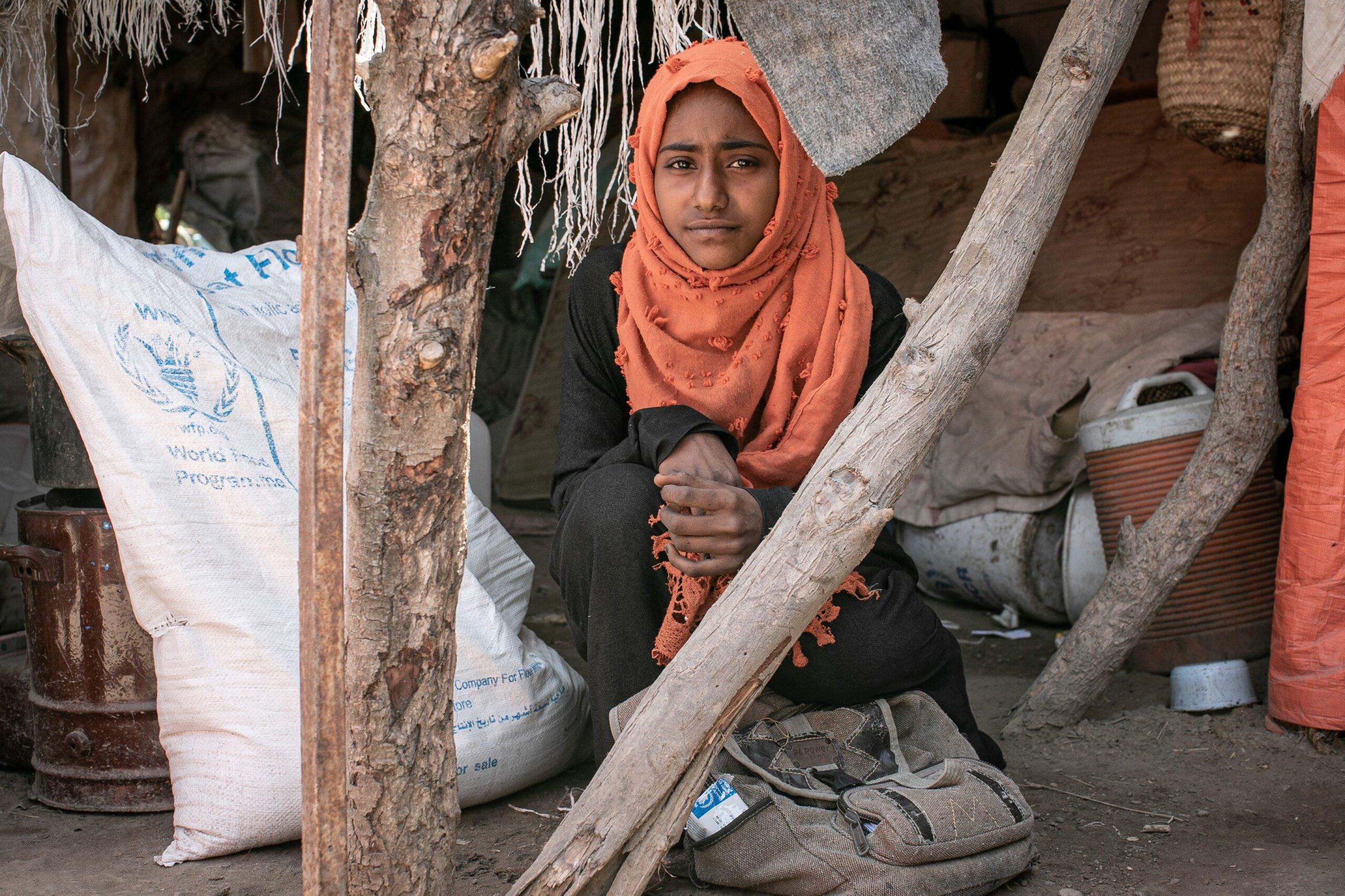 A young Yemeni girl in an orange hijab sits in a makeshift shelter, surrounded by sacks of World Food Program flour, reflecting the urgent need for humanitarian aid. Crypto donations via LTC offer a direct path to provide food security, health, and water, alleviating suffering in war-torn Yemen.