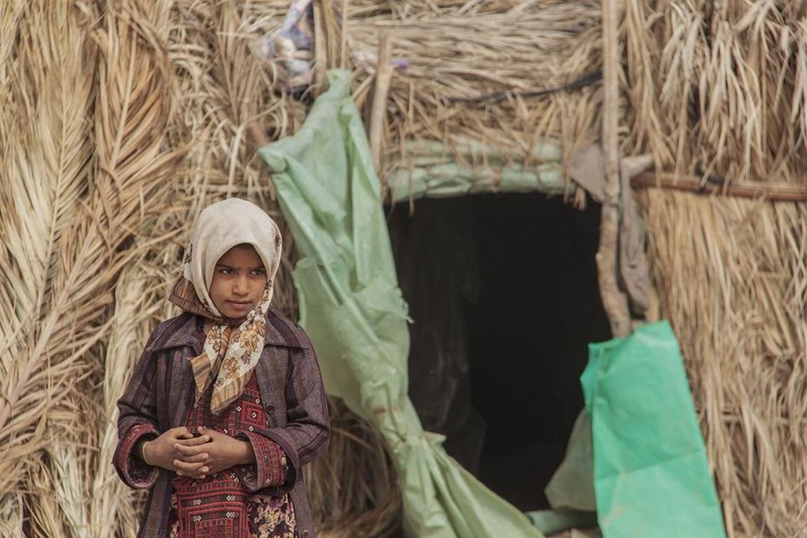 Zakat online with XRP supports a young girl standing in front of a straw shelter in a refugee camp.