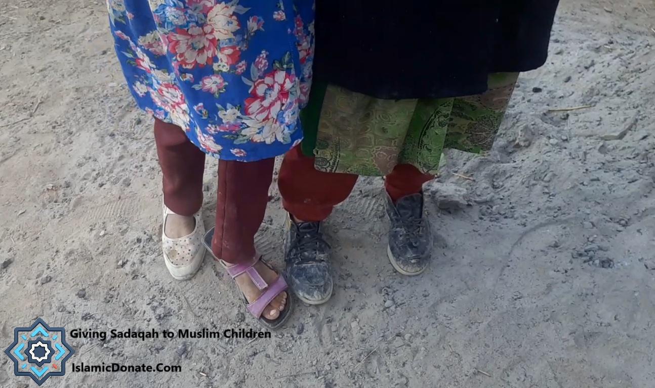 Close-up of children's feet wearing various shoes and sandals, standing on dusty ground. The image depicts the concept of giving Sadaqah, with a focus on providing aid and relief to Muslim children. Your donations, made via RLUSD, support essential needs like meals and family relief.