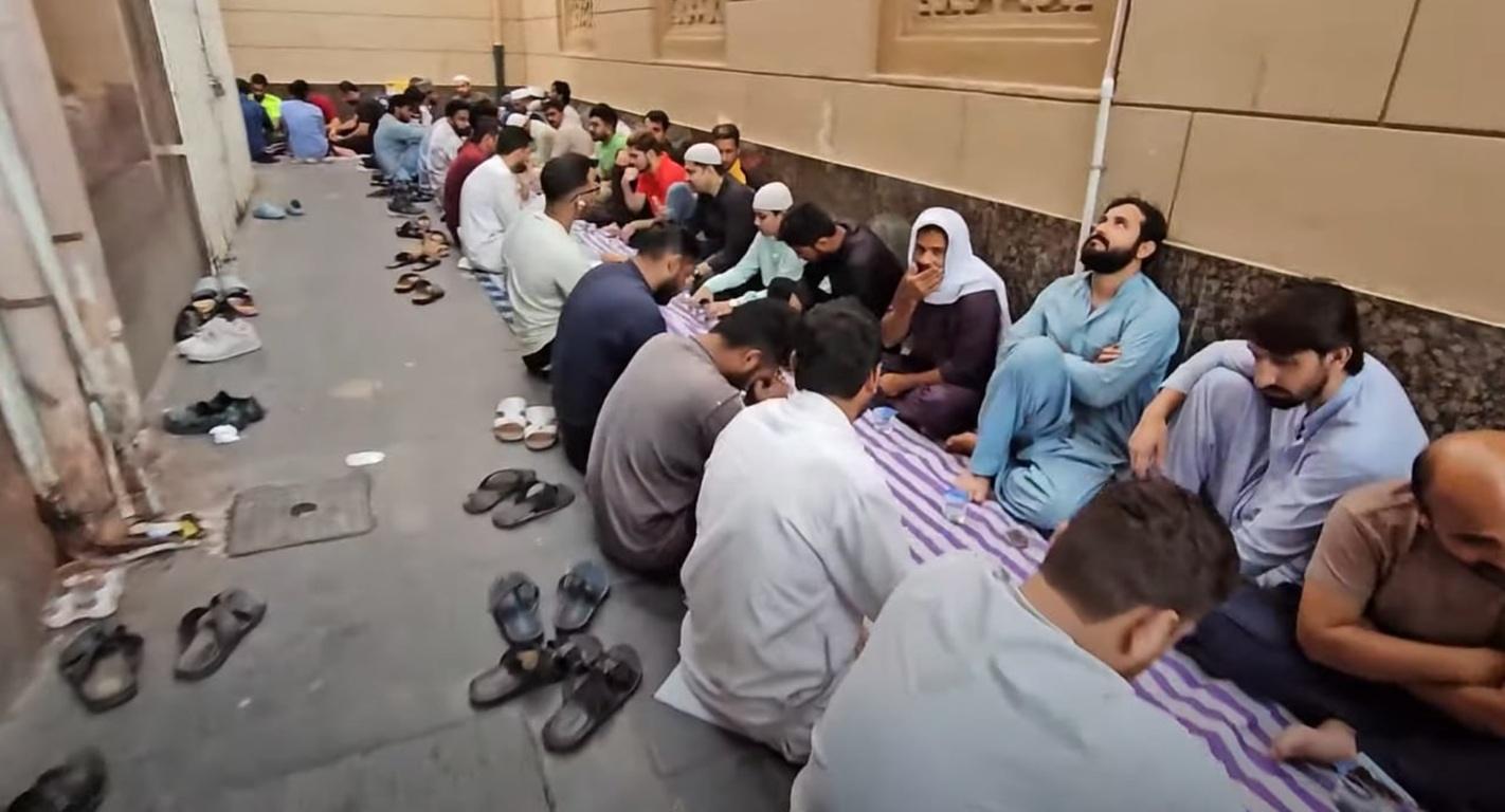 A group of Muslim men sitting on striped mats, some looking upwards, gathered for a meal. They appear to be in a communal setting, with shoes nearby. Donations like these can be made easily with cryptocurrency, such as BTC, for Qurbani and Islamic charity.