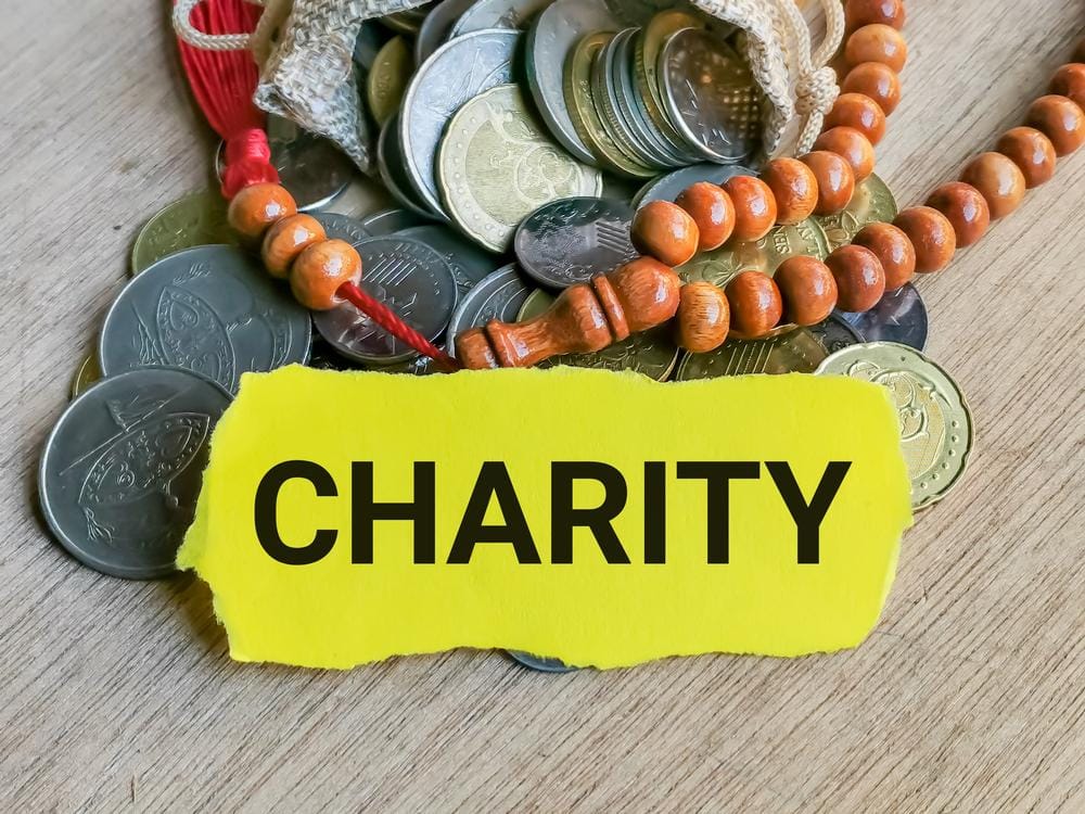 Coins and prayer beads with a yellow slip of paper reading CHARITY, representing Muslim aid and Zakat donations, possibly processed via ETH.