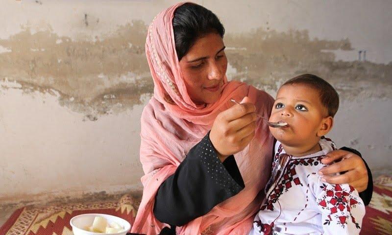 A woman in a pink headscarf feeds a baby from a spoon, supporting Islamic relief efforts via anonymous crypto donations, potentially using BTC for processing.