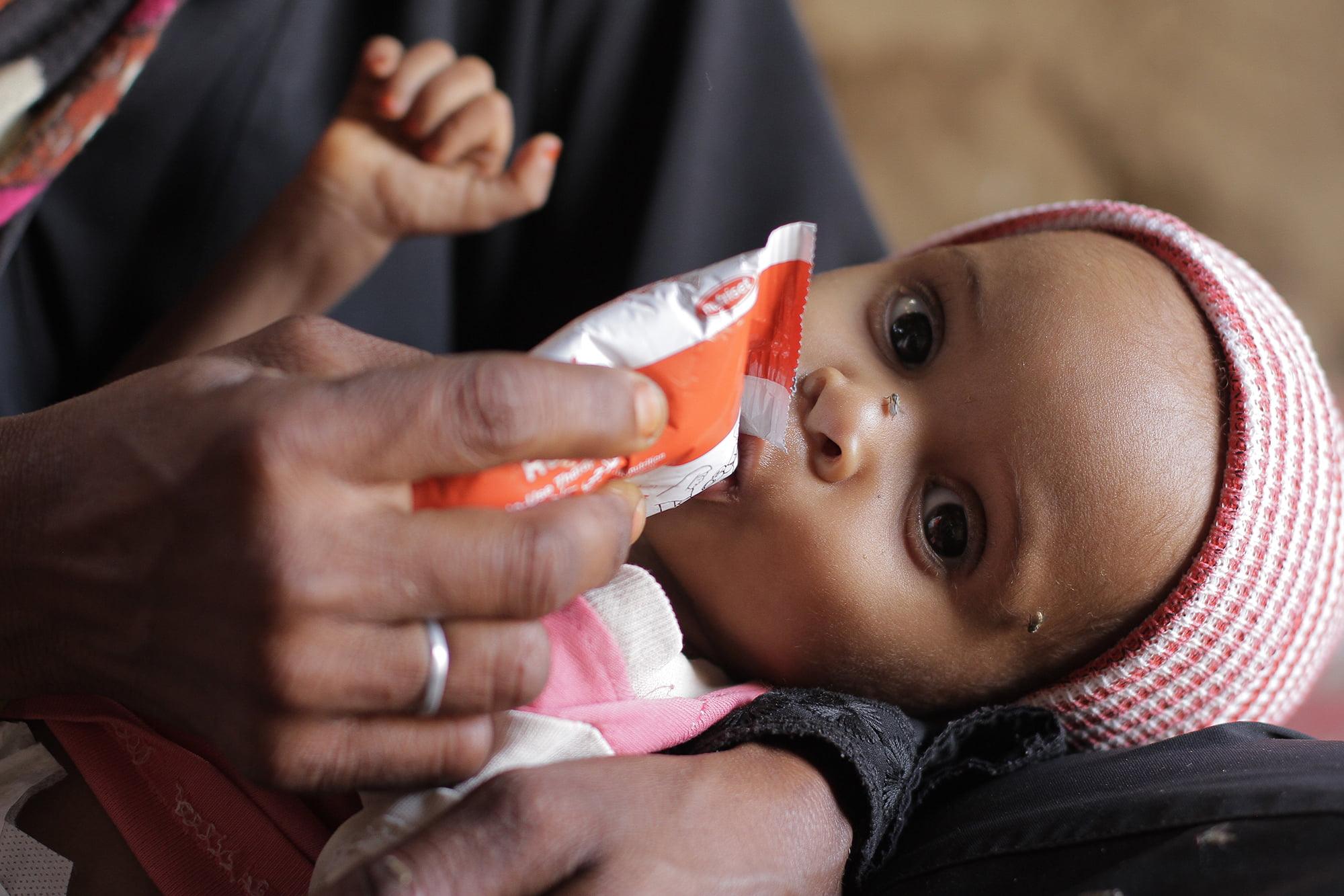 A person is feeding a baby with a packet of food, implying crypto-supported aid. The baby wears a red and white striped hat.