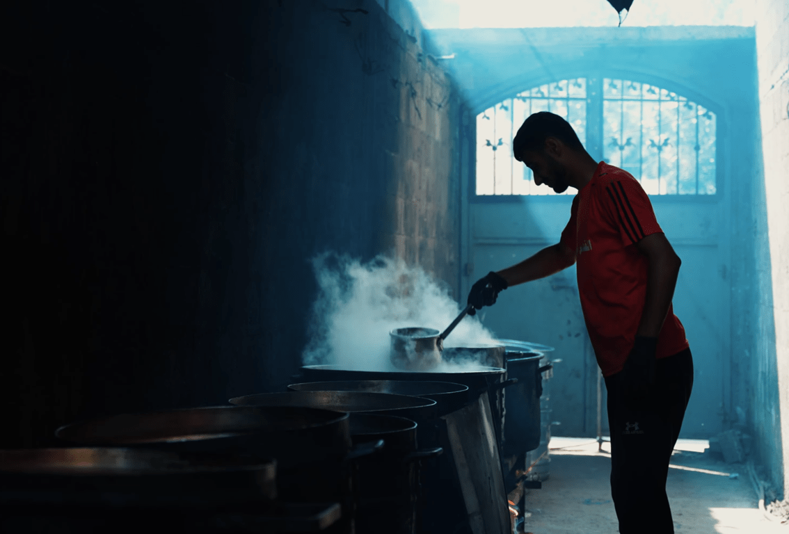 Man stirring pots of food in smoky conditions, symbolizing communal kitchens and aid distribution. Crypto donation supports immediate relief for displaced families in Palestine. ETH accepted.