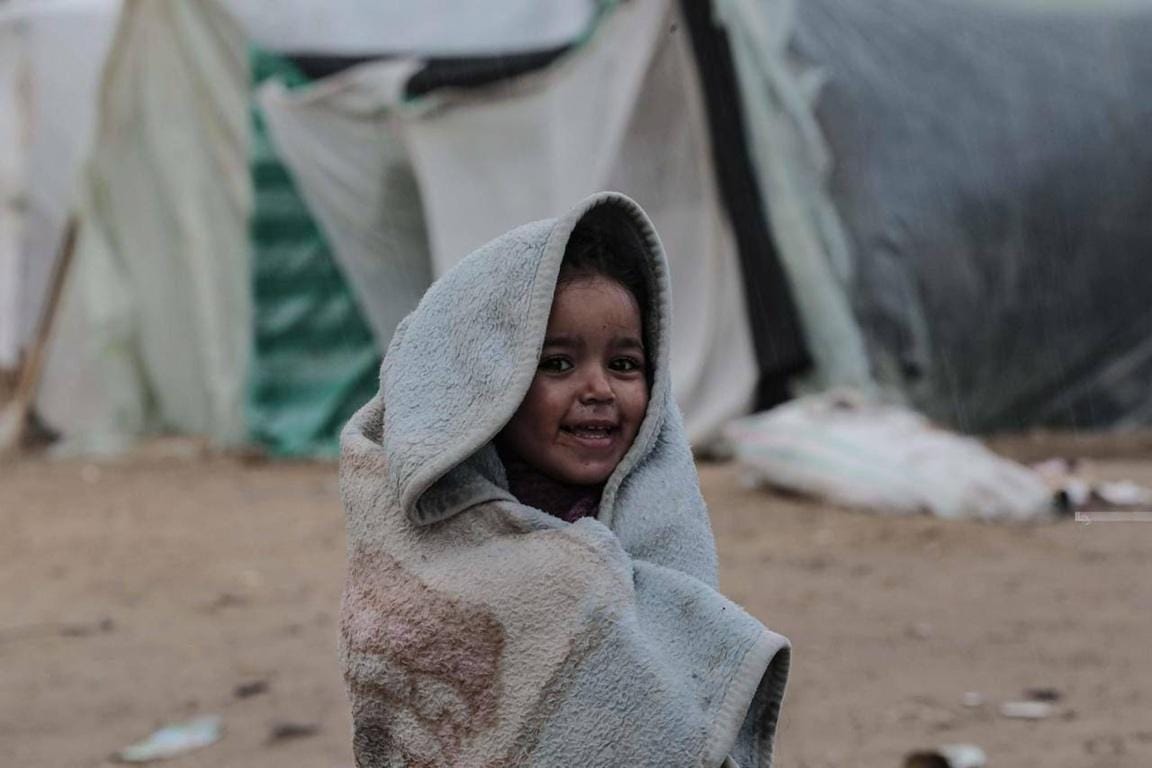 Smiling child wrapped in a blanket in a camp, representing IslamicDonate's Aqiqah Qurbani service, made possible with BTC donations.