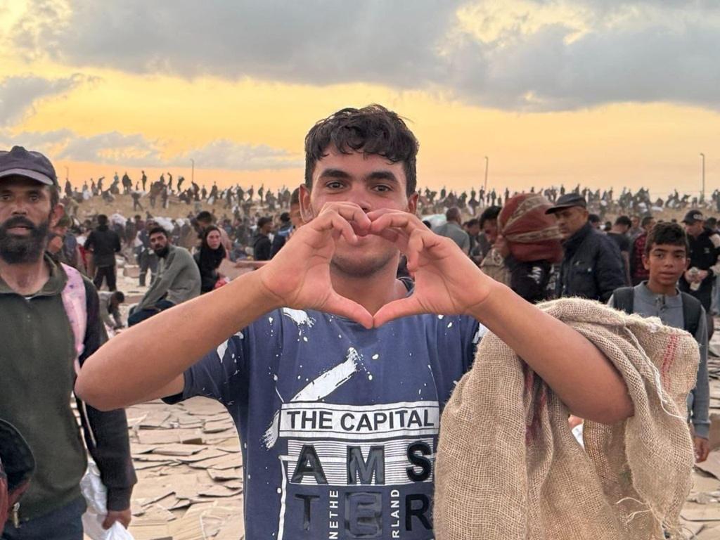 Young Palestinian man forms a heart shape with his hands, symbolizing hope and resilience amidst a crowd of people in a damaged area. Your crypto Zakat, like Stablecoin, can provide vital aid for food, water, and reconstruction efforts.