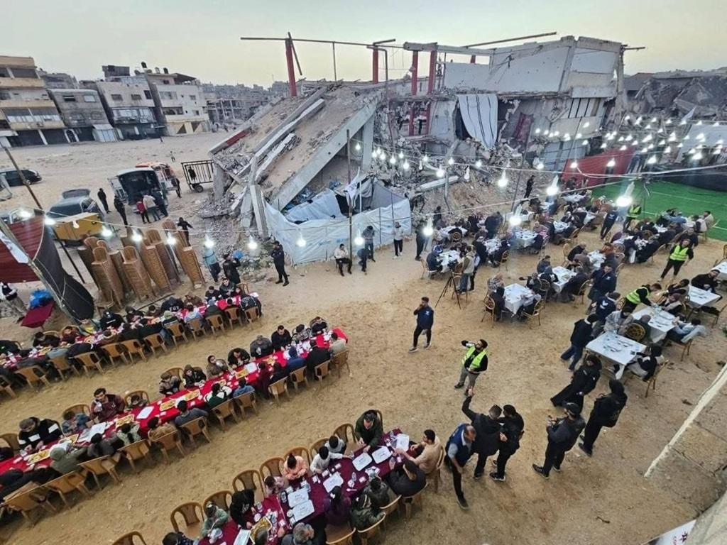 Ramadan Iftar tables are set amidst destroyed buildings in Gaza. People sharing meals under string lights. Aid for Palestinian fasters, donate via Bitcoin.
