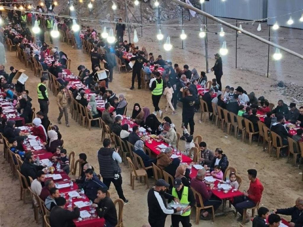 Ramadan 2026 Iftar for displaced families in Gaza. Aid workers distribute meals at long tables set up outdoors under fairy lights. Donations in Ethereum support this vital sustenance for fasting believers.