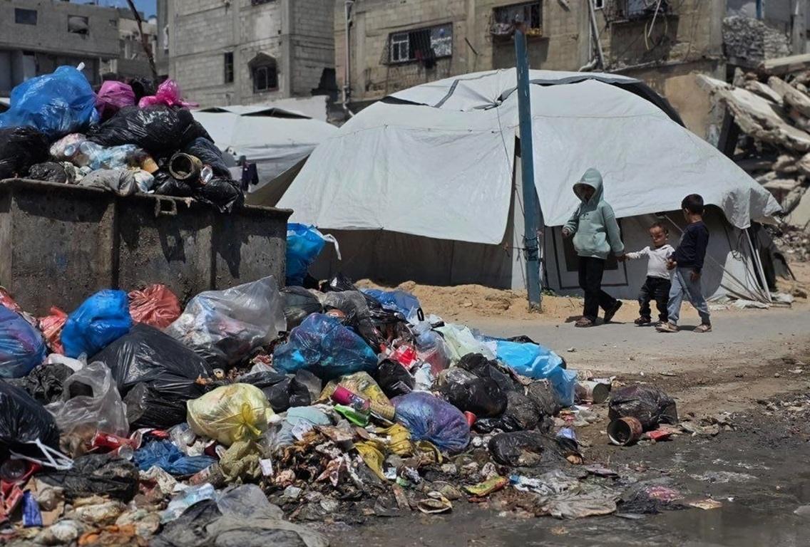 Gaza summer hazards show children walking near rotting garbage and tents. Send USDC to fund hygiene kits and clean water.