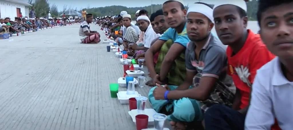 Ramadan food assistance distribution to fasting individuals, supported by crypto donations like ETH. The scene shows many people sitting in rows, with food containers and cups laid out before them, representing a relief effort.