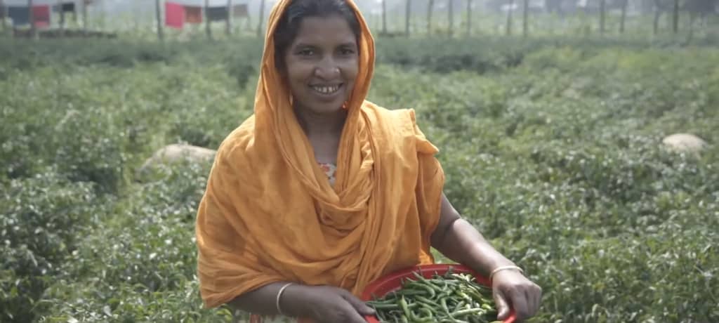 Aisha, an empowered woman in rural Bangladesh, holds a basket of fresh crops signifying the success of her farm, made possible by crypto donations and community support. Her story exemplifies how empowering women leads to flourishing families. Donation via XRP.