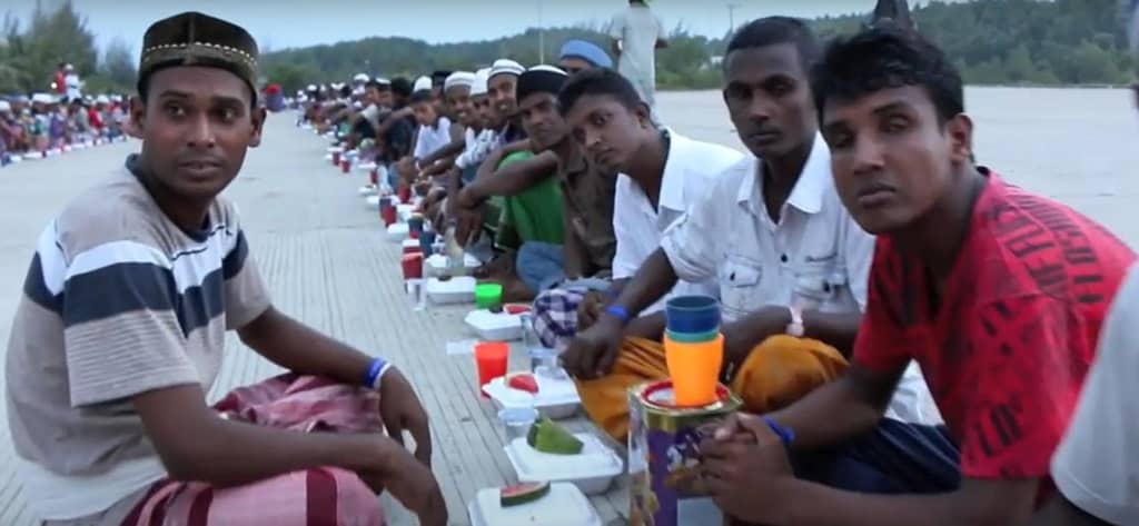 Men sit in rows receiving food aid, with packaged meals and drinks arranged before them. The scene shows the impact of humanitarian efforts, including crypto donations like TRX, supporting those in need of suhoor and iftar.
