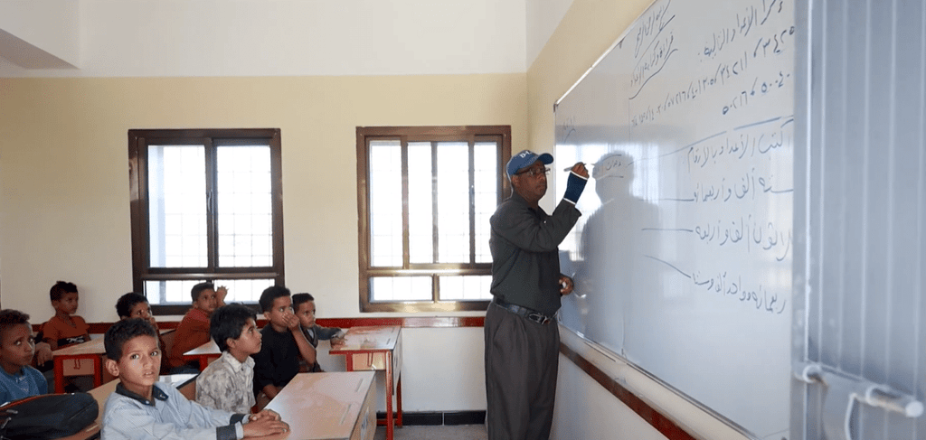 Sponsor of orphans classroom with teacher writing on whiteboard, crypto donations enable education for 49 children sponsored via ETH.
