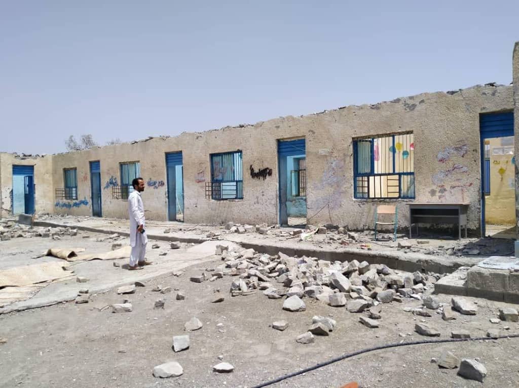 Man stands amidst rubble outside a damaged school building, symbolizing the need to donate for education with crypto, possibly via USDC, to support children's access to learning.