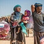 Family carrying donated blankets and relief supplies in a dusty, arid village, aided by crypto, likely for winter relief.