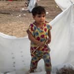 A young child stands amidst makeshift shelter fabric, symbolizing the ongoing daily struggles and resilience in Palestine. Aid delivery is hampered by checkpoints, waste management issues, and water scarcity. Supporting this cause with BTC donations helps overcome obstacles in delivering essential resources and education.