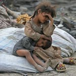 Two children huddled together amidst debris, one older sibling comforting the younger. Digital donations through SOL support aid.