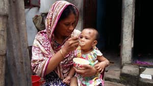 A woman feeds a baby from a bowl, likely a scene of charitable aid supported by USDT, illustrating humanitarian relief.
