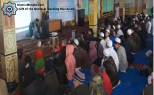 Children attending a Quranic education session, learning vital Islamic teachings, with one teacher presenting via a projector, supported by cryptocurrency donations like BTC.