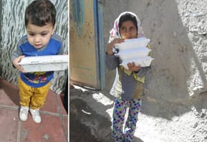 Two children, a young boy and a girl, hold containers of food. The boy wears a blue and grey shirt and yellow pants, carrying one container. The girl, in a headscarf and colorful pants, carries a stack of three containers. Serving food to the needy with ETH donations.