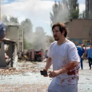Bloody medical professional with phone in hand walks through debris after missile attack on Kyiv children's hospital, seeking aid via BTC donations.