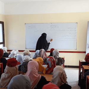 Classroom of young girls in headscarves learning from a teacher writing on a whiteboard, supported by crypto donations like ETH.