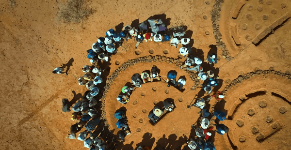 Aerial view of a diverse group of people gathered in a circular formation, participating in a tree planting or reforestation project in a dry, arid landscape. Donations support environmental protection initiatives, including reforestation efforts, with a nod to crypto donations like SOL.