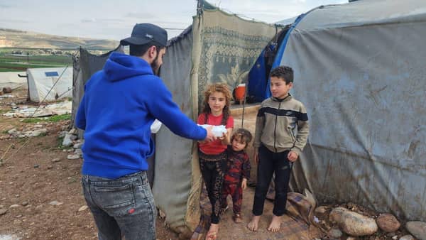 A volunteer distributes aid to children near tents in Aleppo, with support facilitated by USDT. Aid includes food, medicine, shelter, and water, embodying Islamic principles of charity. Keywords: Aleppo crisis, humanitarian aid, crypto donations, Islamic charity, poverty and hunger alleviation.