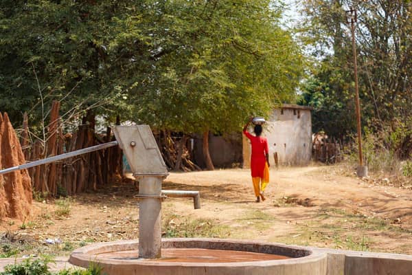 A rural scene with a water pump in the foreground and a woman carrying a pot on her head in the background. Donate clean water via USDT for Islamic charity.