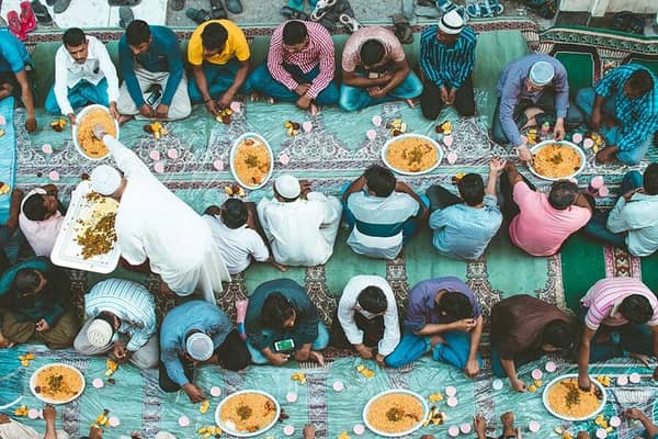 A group of men sit on the floor eating from large platters of rice, with a man serving them from a large tray. This scene illustrates Muslim food culture and ethical eating, with donations facilitated by SOL.
