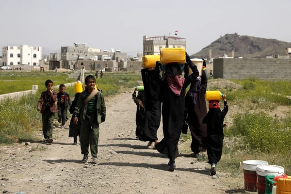 Children and women carrying water containers on their heads in a dusty village, with buildings in the background. Aid supported by Arab Emirates Dirham.
