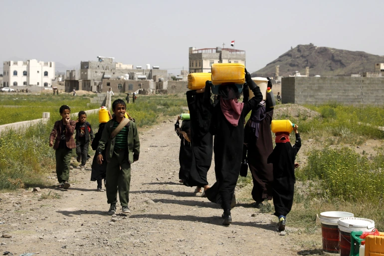 Children and women carrying water containers on their heads in a dusty village, with buildings in the background. Aid supported by Arab Emirates Dirham.