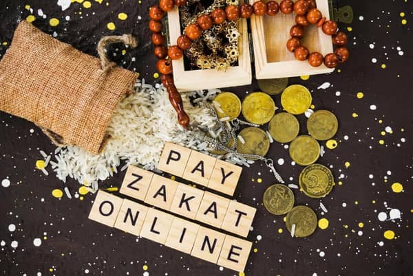 Text reads PAY ZAKAT ONLINE. Islamic prayer beads, gold coins, and a burlap sack of rice are scattered on a dark, speckled background.