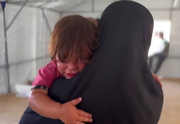Close-up of a crying child being held by a mother wearing a black hijab. The child's face is wet with tears, and their hand rests on the mother's shoulder. Urgent need for aid in Palestine, with donations via BTC.
