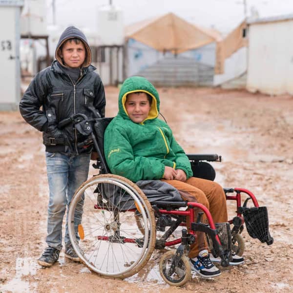 A boy in a wheelchair smiles next to his friend in a refugee camp, receiving aid via BTC.