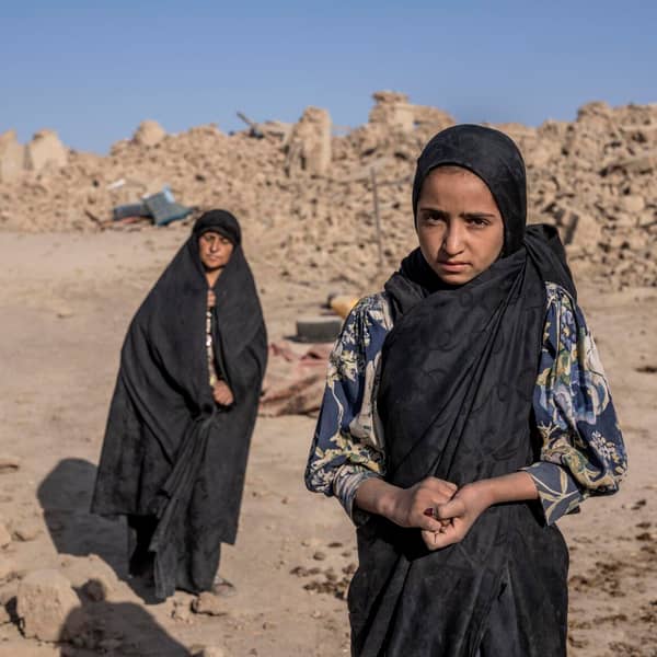 Two Afghan women stand in rubble after the 2023 earthquake, one looking at the camera with a serious expression. Providing first aid kits, training, and supplies, crypto-enabled donations like USDT support relief efforts for earthquake victims.