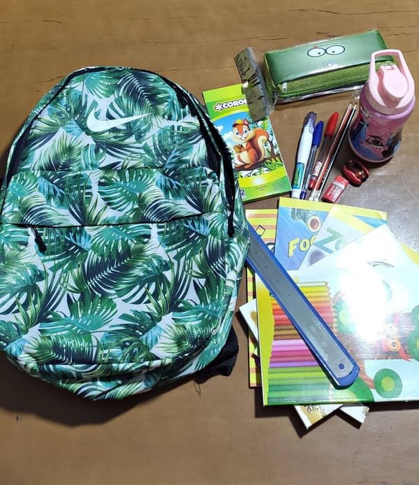 A vibrant green backpack with a tropical leaf pattern is displayed alongside school supplies including notebooks, pens, pencils, a ruler, and a water bottle. This image represents a donation for education, facilitating children's back-to-school needs, with support processed via USDT.